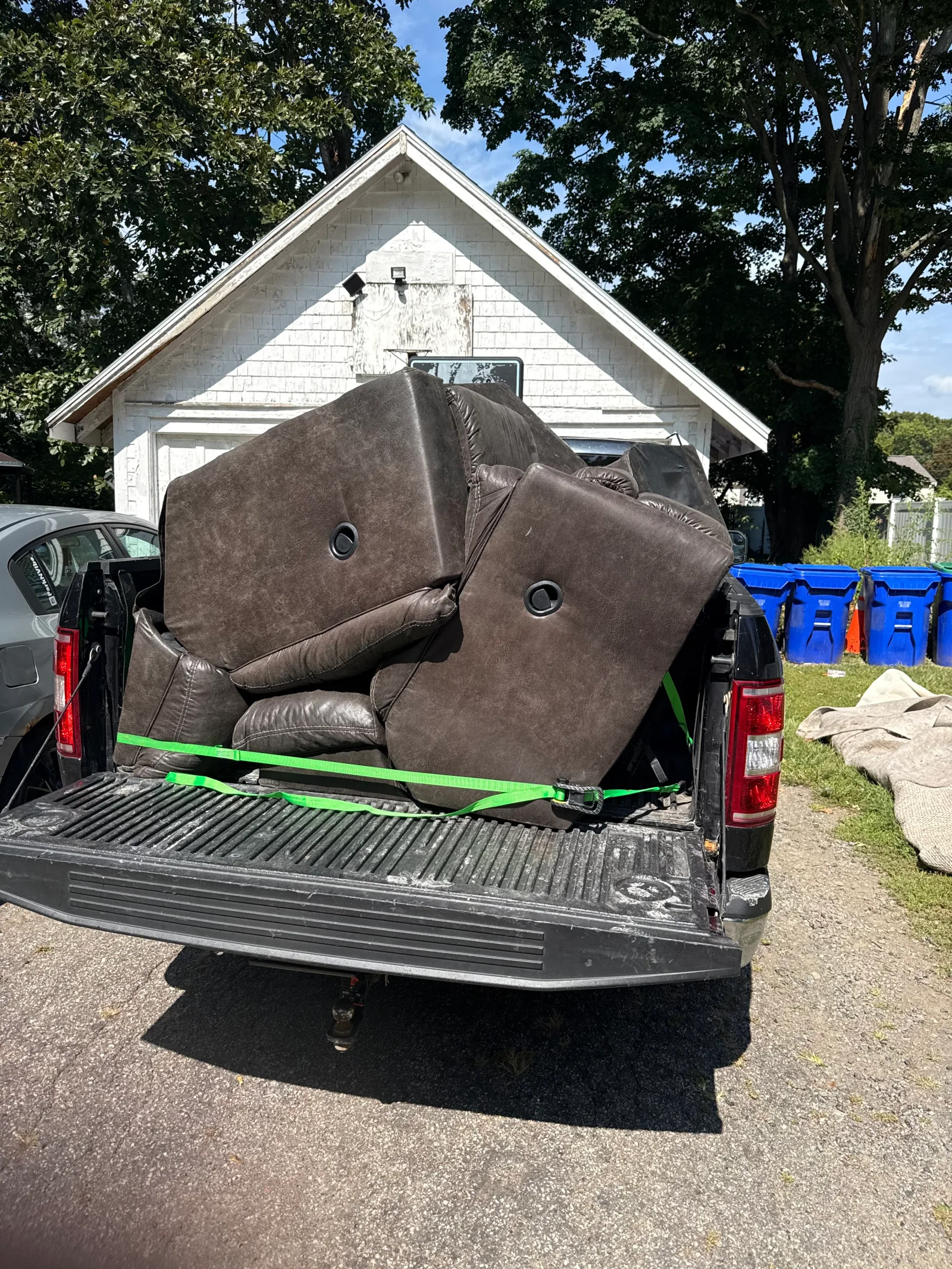 Dark brown faux-leather sofa and recliner cushions strapped in a pickup truck bed with green ratchet straps for junk removal