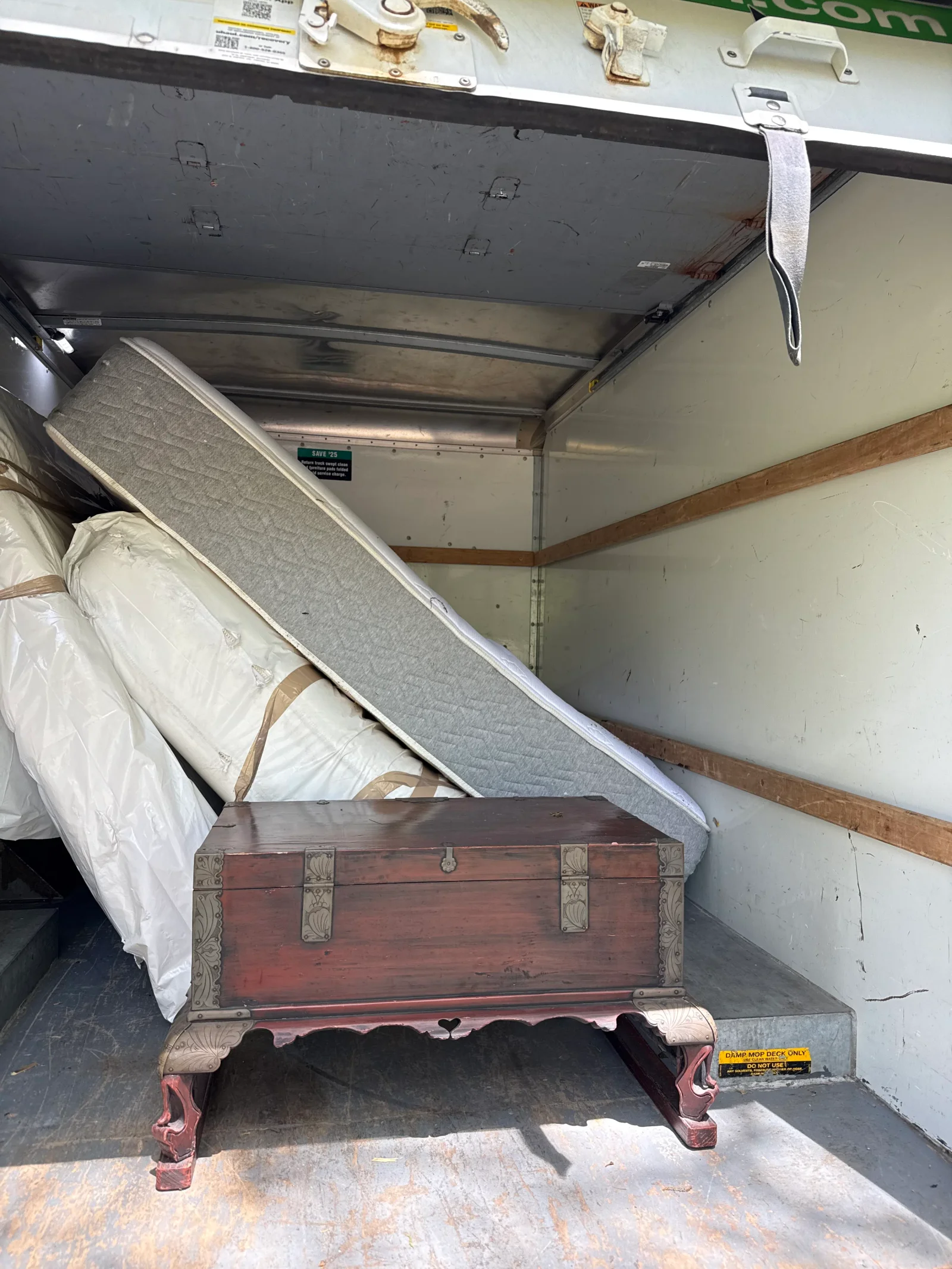 Box truck interior with wrapped mattresses strapped upright and an ornate wooden chest with metal fittings on the floor