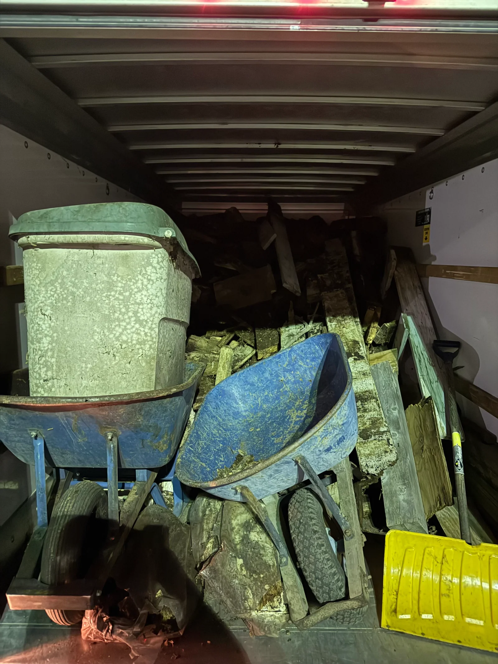 Box truck interior filled with blue wheelbarrows, weathered wood planks, a gray plastic bin, and a yellow shovel