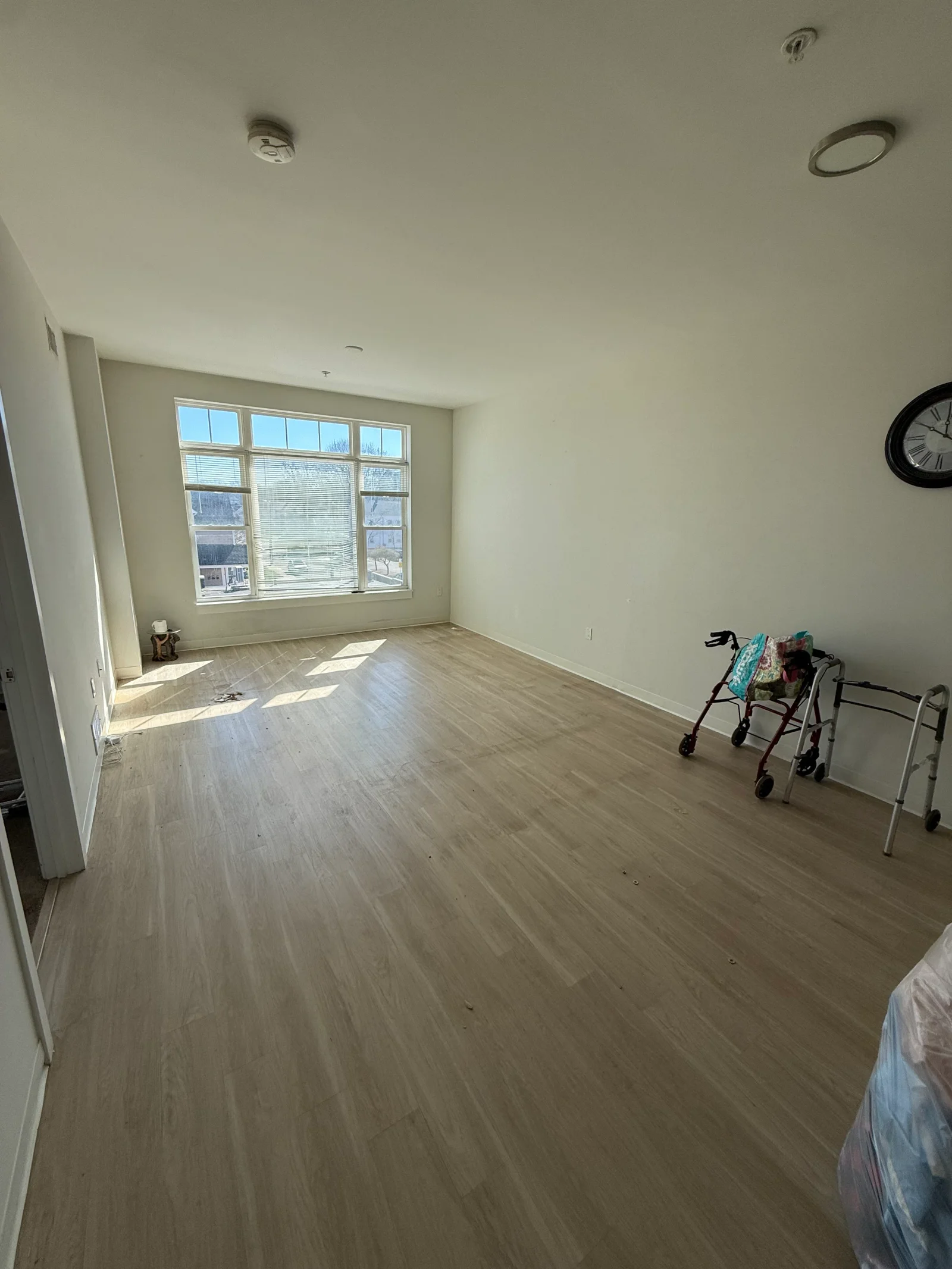 Empty living room with light wood-look vinyl plank flooring, white walls, and large window with blinds after clearing