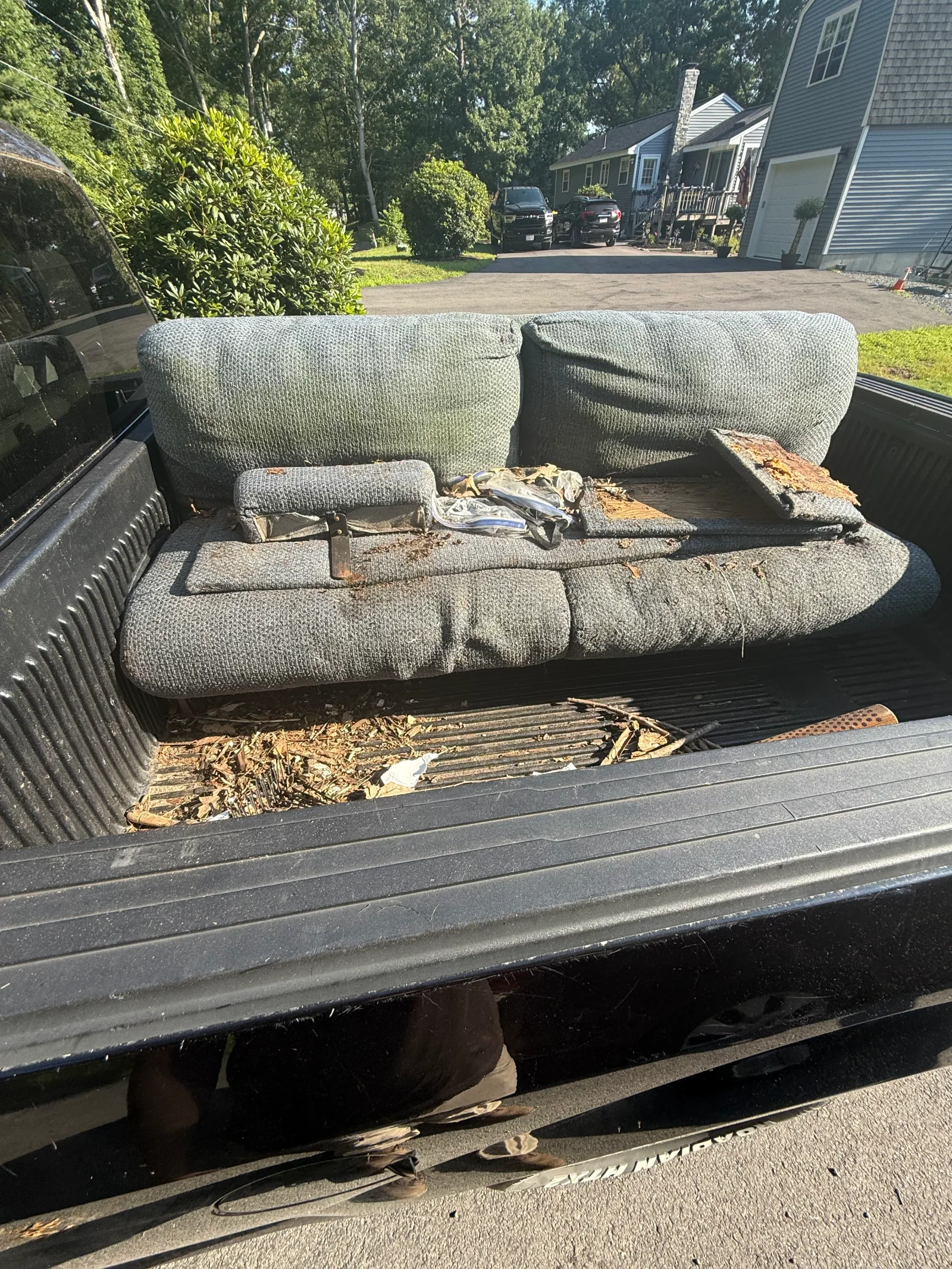 Worn gray upholstered sofa with torn fabric and exposed foam loaded in a black pickup truck bed with debris