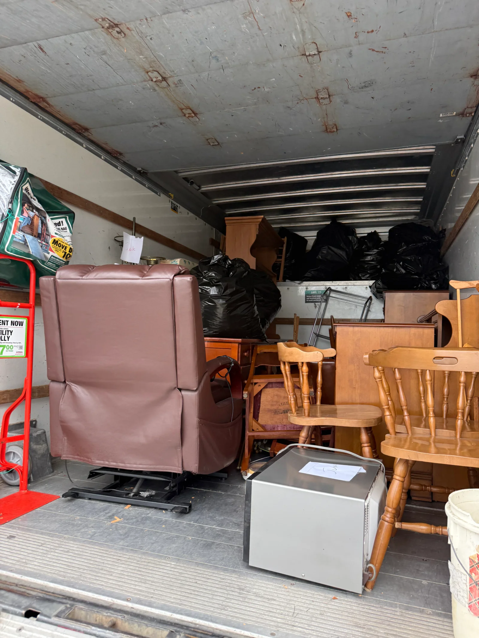 Box truck interior loaded with brown recliner, wooden chairs, cabinet, microwave, and black trash bags