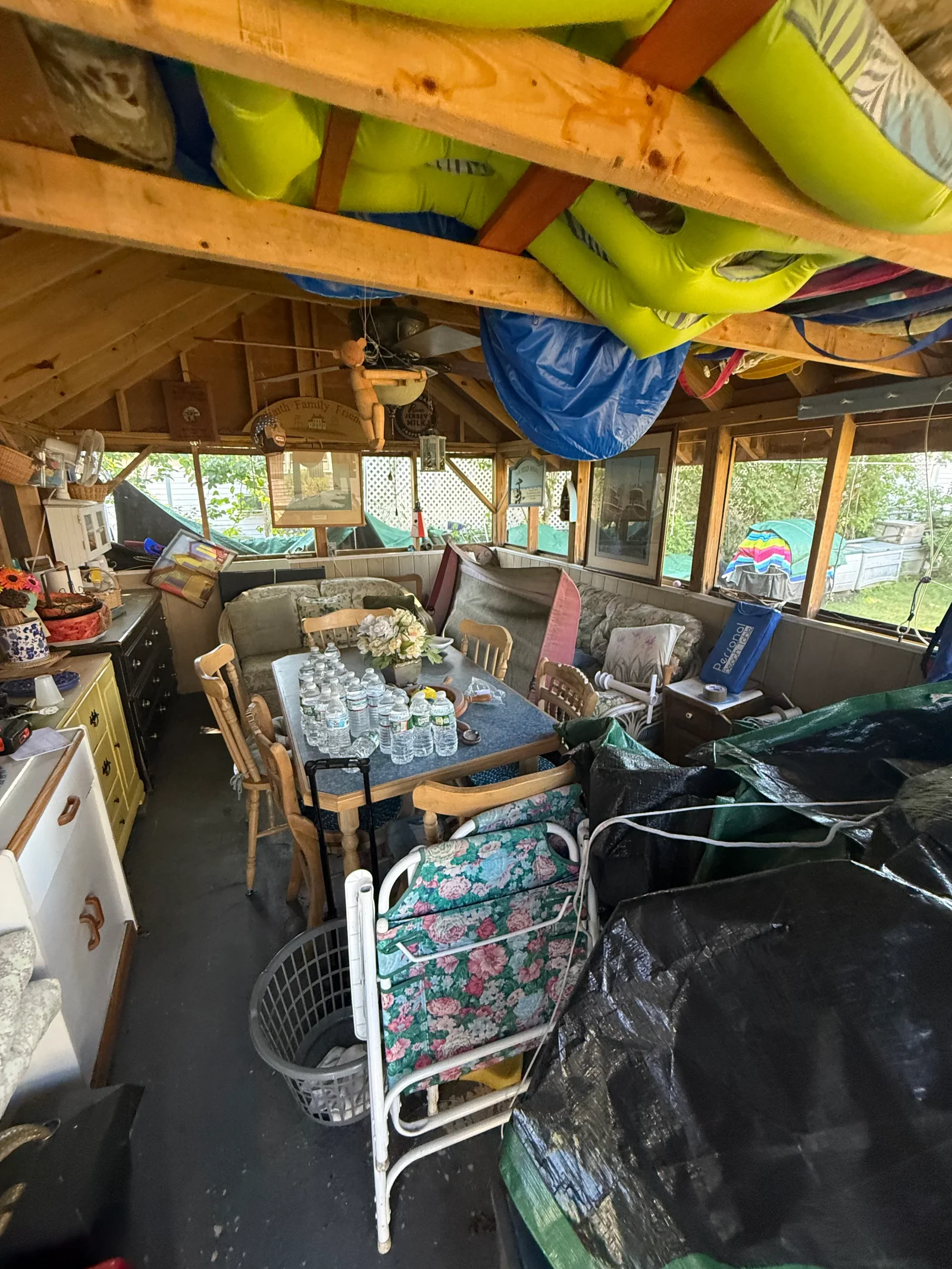 Wood-framed sunroom interior filled with inflatable pool toys on exposed rafters, upholstered chairs, wooden dining chairs, and stored tarps before junk removal.