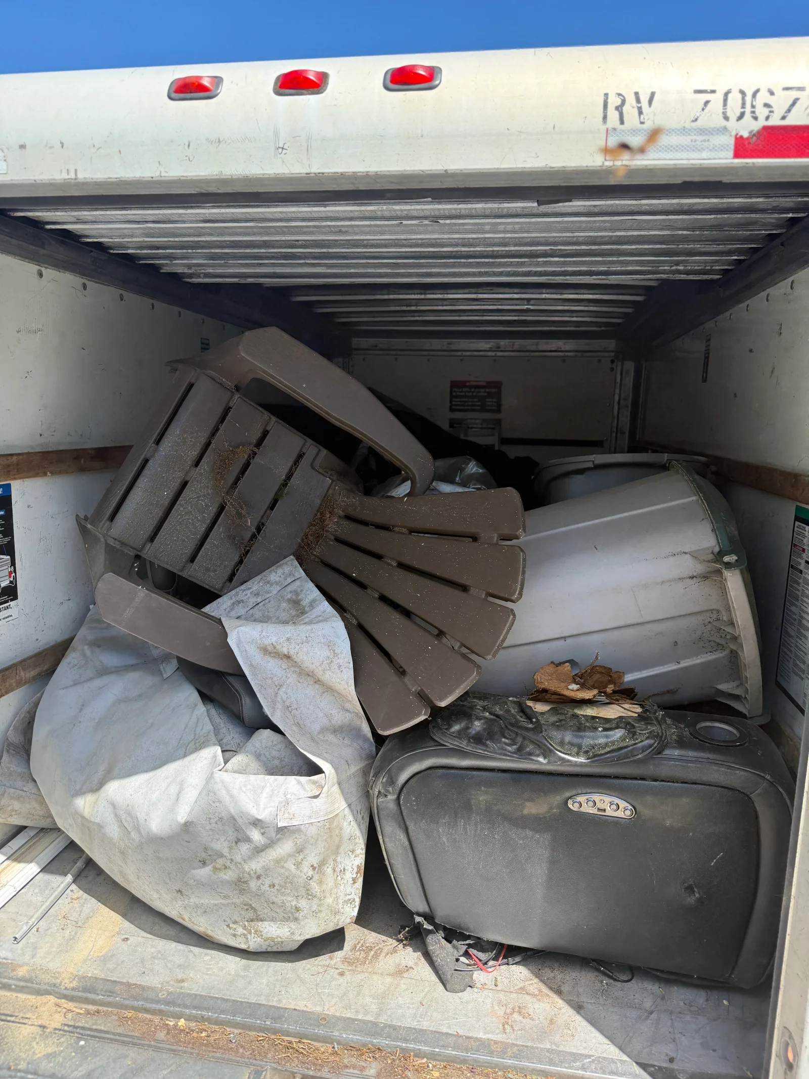 Box truck interior loaded with brown plastic chair, gray plastic bin, black vinyl seat, and dirty white tarp for junk removal