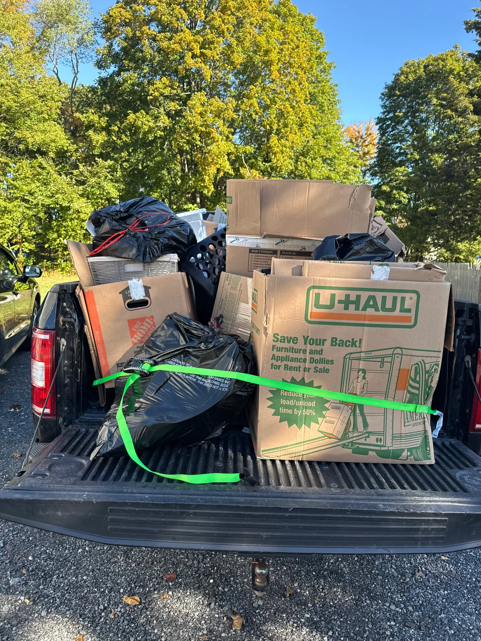 Pickup truck bed loaded with cardboard boxes, black trash bags, and a laundry basket secured with a green ratchet strap