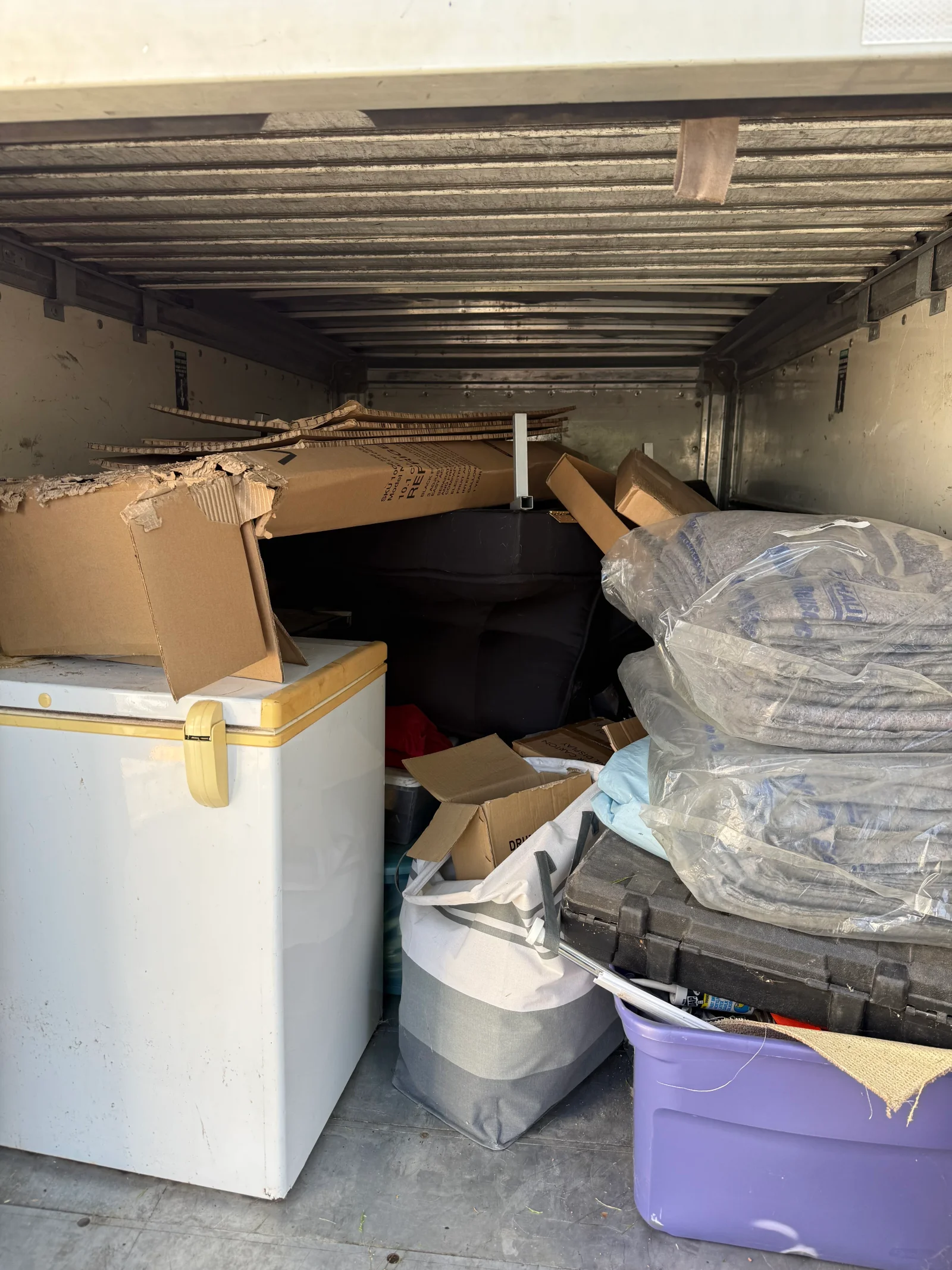 Box truck cargo area filled with white chest freezer, torn cardboard, bagged gray blankets, purple tote, and black equipment case