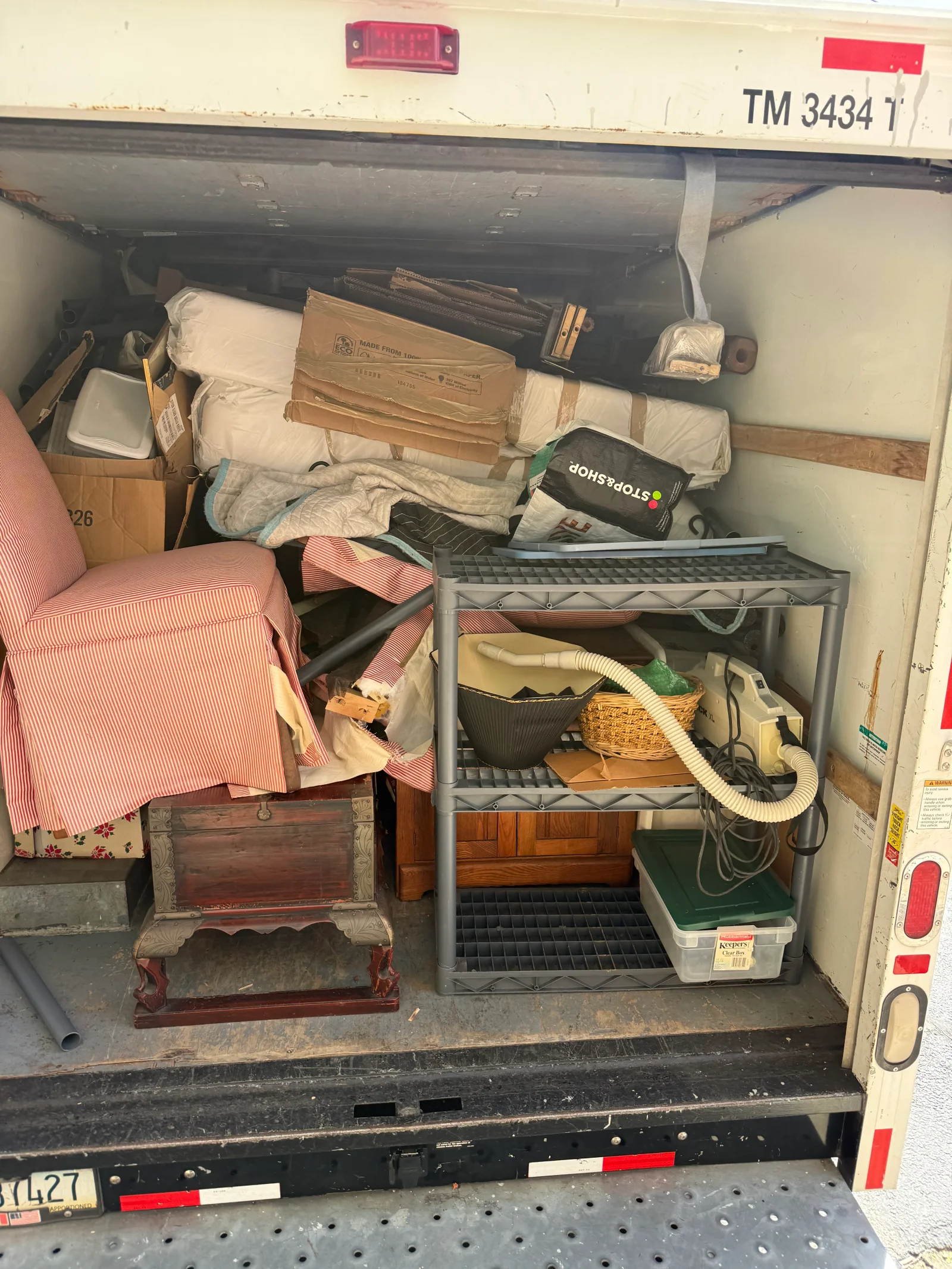 Box truck cargo area filled with striped armchair, wooden chest, wrapped mattresses, cardboard boxes, and wire shelving unit