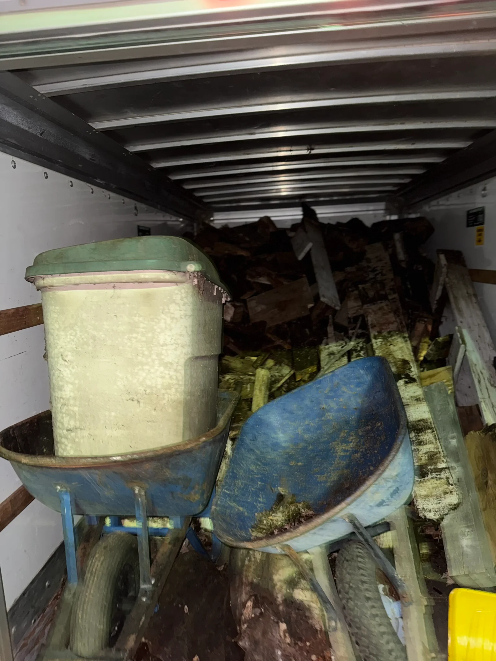 Enclosed box truck loaded with broken wood, debris, and two blue wheelbarrows, one holding a cream plastic container
