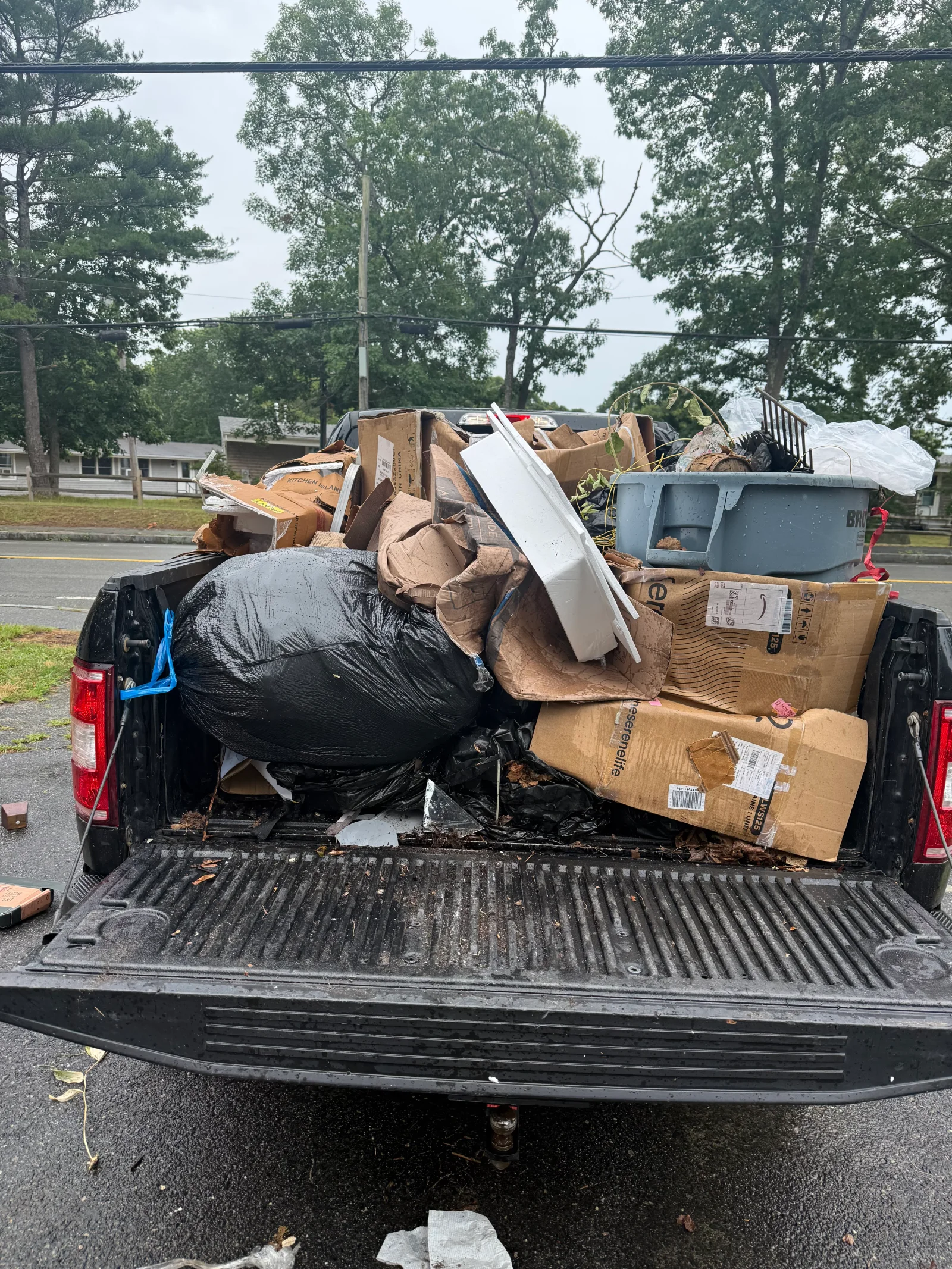 Pickup truck bed loaded with cardboard, black trash bag, foam board, plastic tote, and yard debris for junk removal