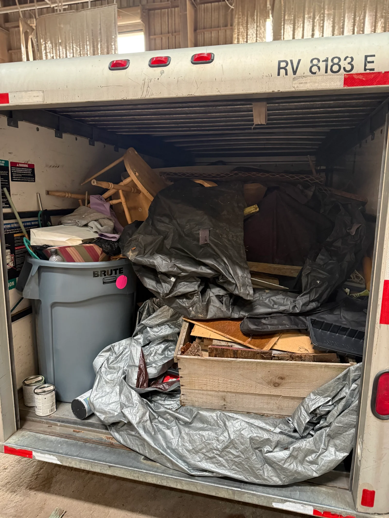 Rental truck cargo area packed with wooden crate of scrap lumber, tarps, broken chair, and gray trash can