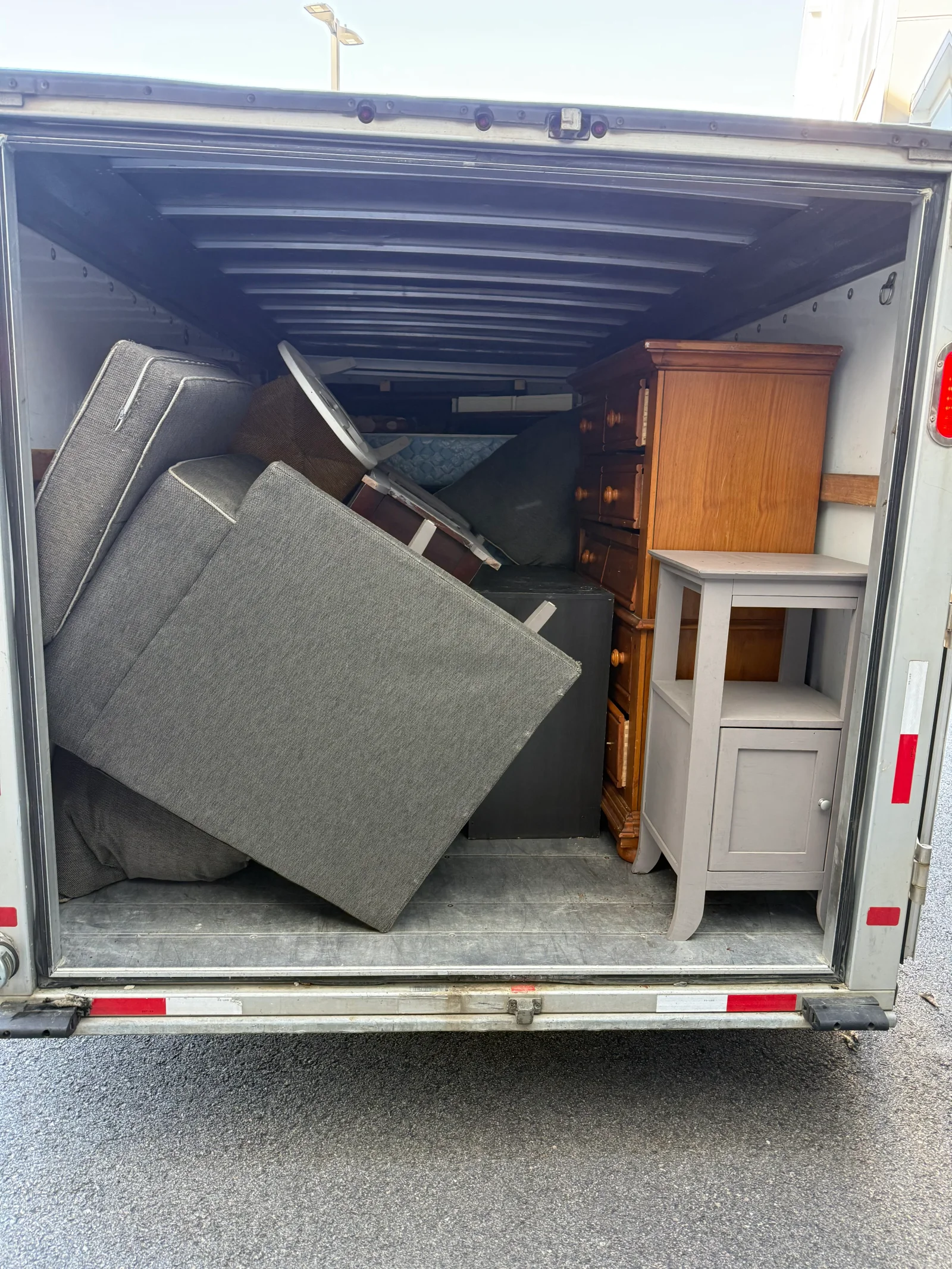 Cargo van interior packed with gray upholstered cushions, a wood dresser, a black cabinet, and a gray side table for junk removal
