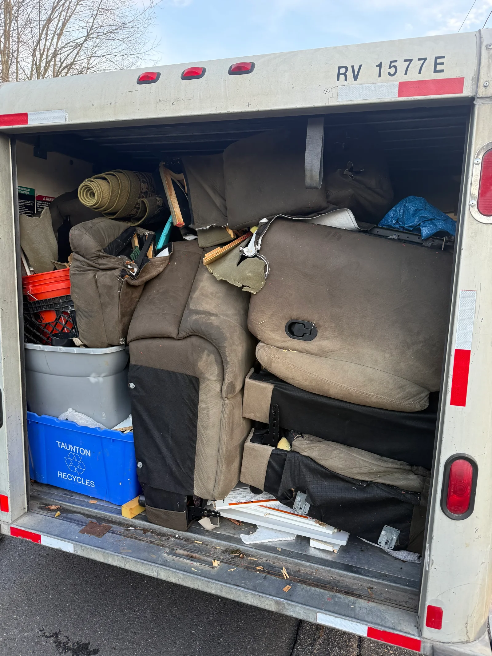 Box truck interior packed with brown upholstered sofa cushions, recliner, rolled carpet, and plastic bins for removal