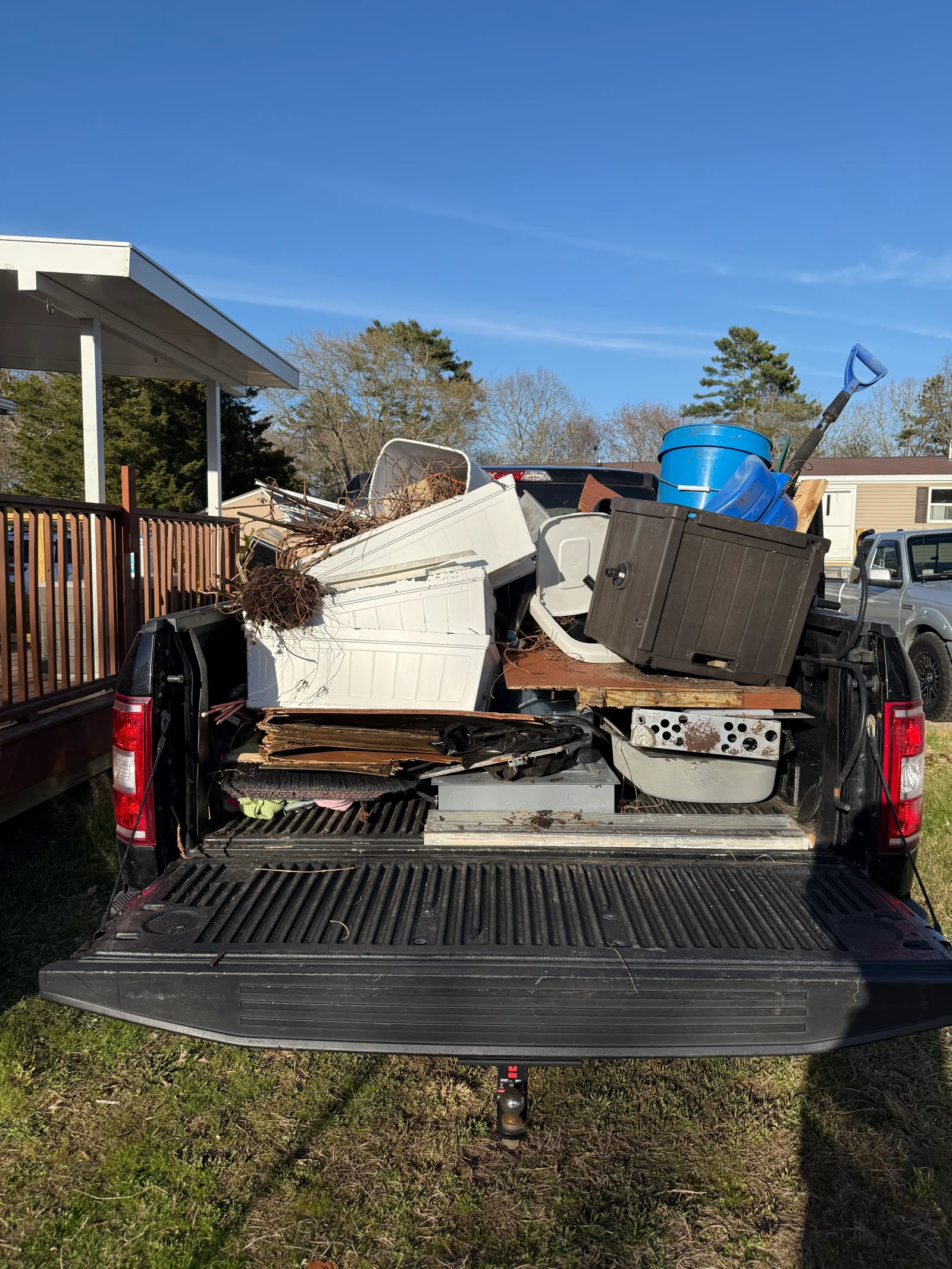 Pickup truck bed packed with plastic bins, wood planks, metal debris, a blue barrel, and dried branches for junk hauling
