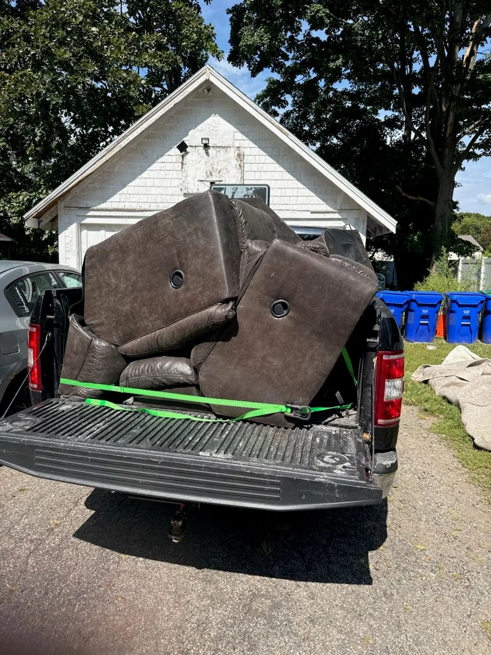 A worn sofa and wooden dresser staged at the curb of a residential home in Taunton, Massachusetts, ready for furniture removal pickup. An HS Junk Removal and Property Services truck is visible in the driveway.